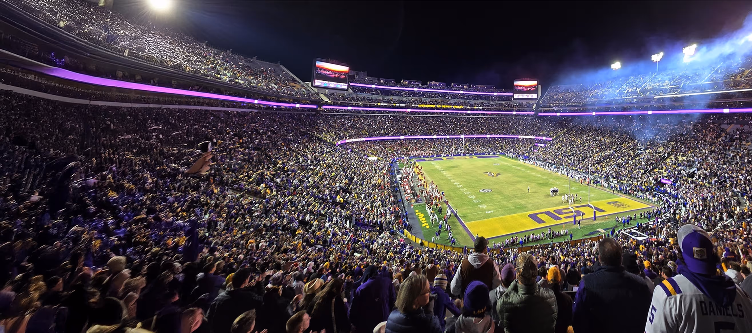Nighttime aerial view of a packed stadium with fans watching a football game on a brightly lit field.