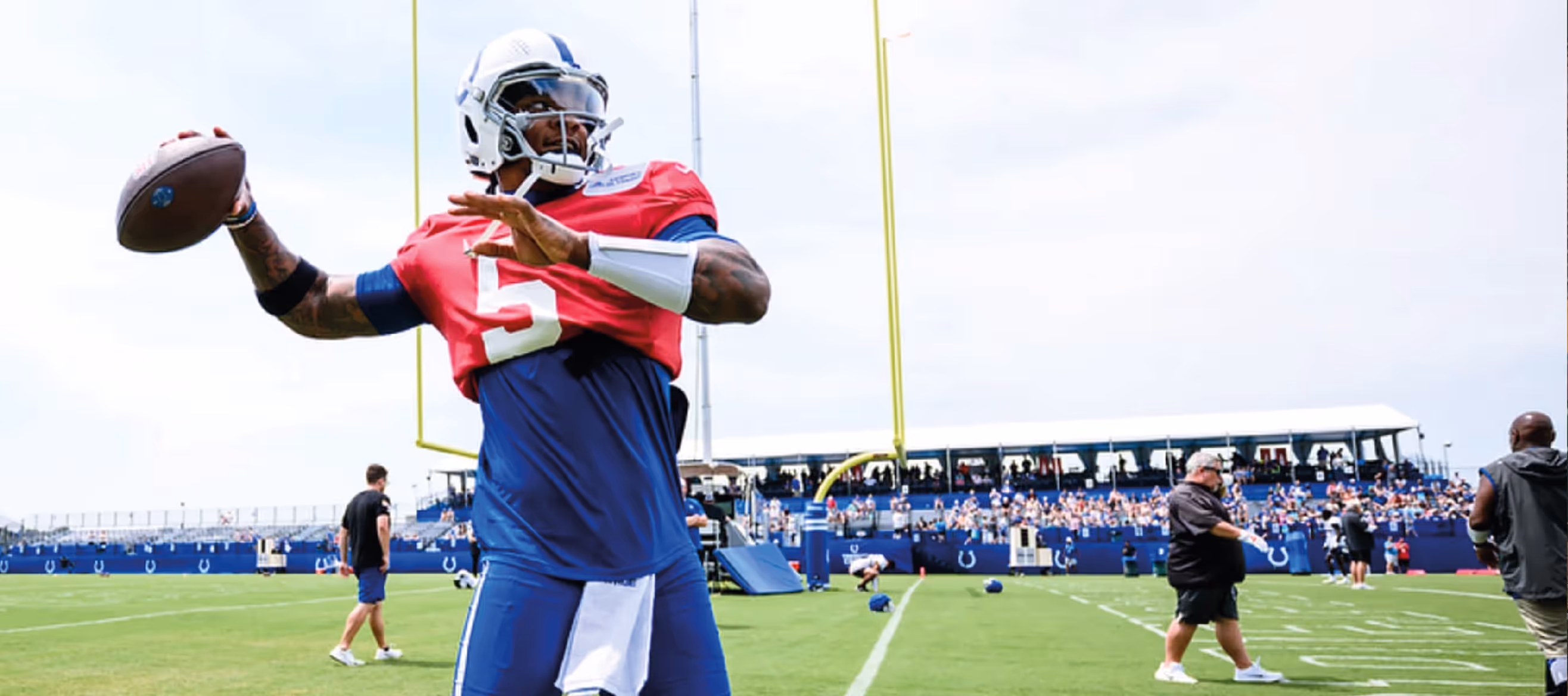 Football player in red practice jersey with number 5 preparing to throw a football on a green field with spectators in the stands.