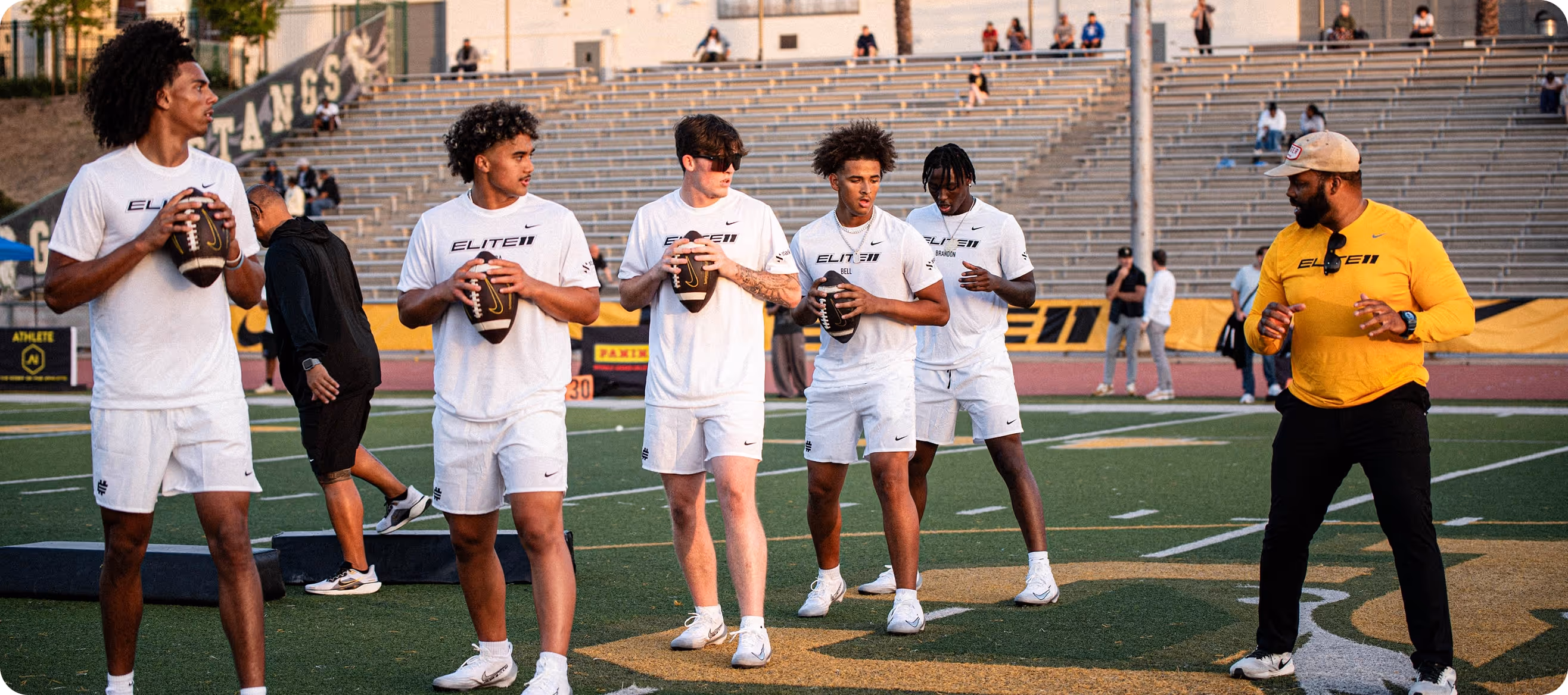 Five young men in white athletic gear holding footballs stand on a field while a coach in a yellow shirt instructs them.