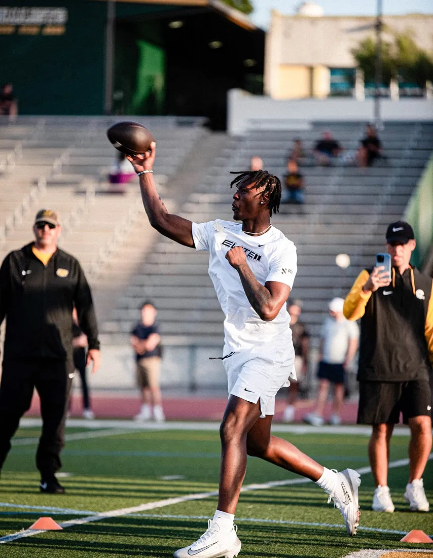 Athlete in white sportswear throwing a football on a field with spectators and coaches in the background.