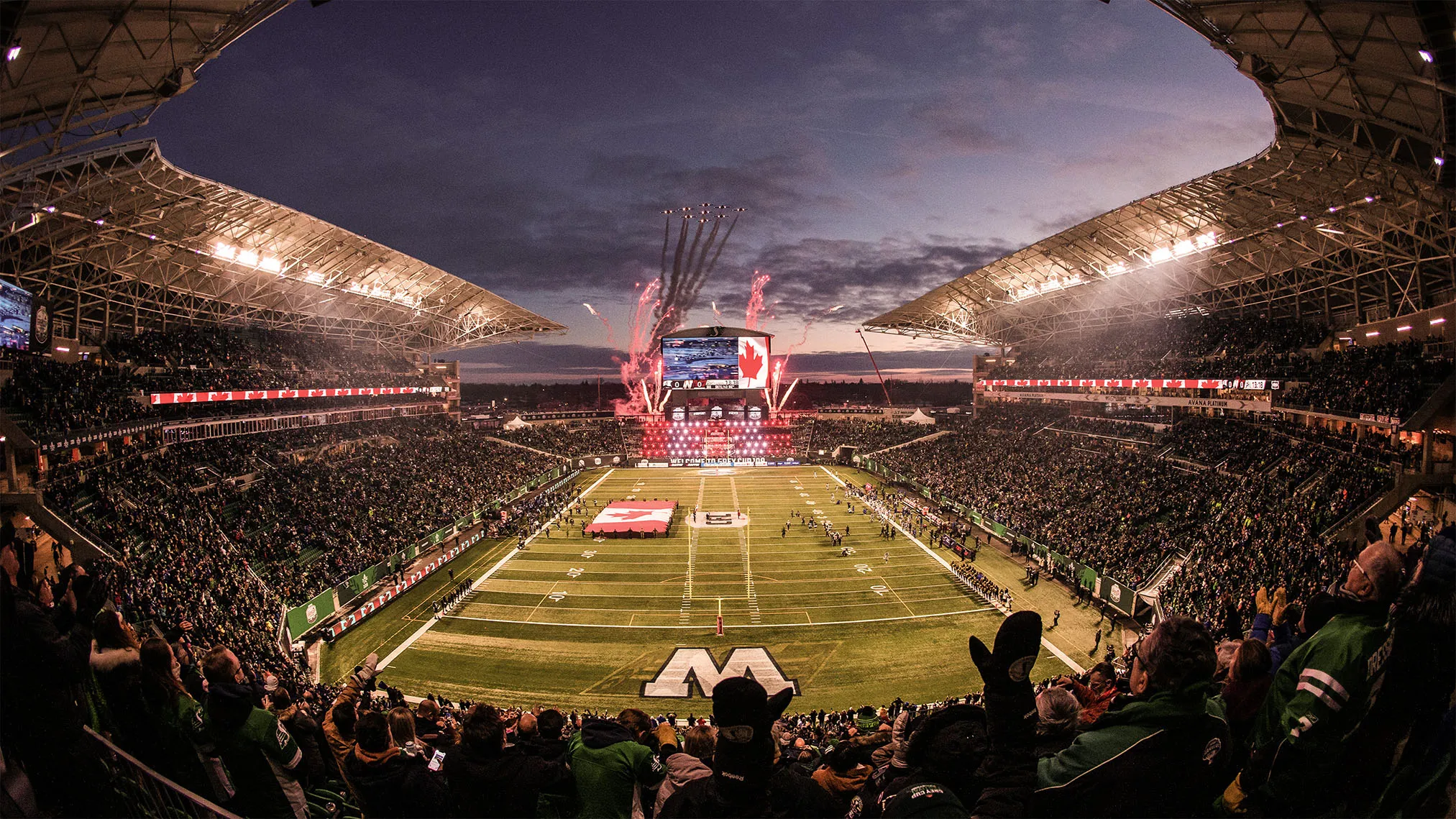 Wide view of a crowded stadium at dusk with Canadian flags, fireworks, and planes flying in formation above the field.