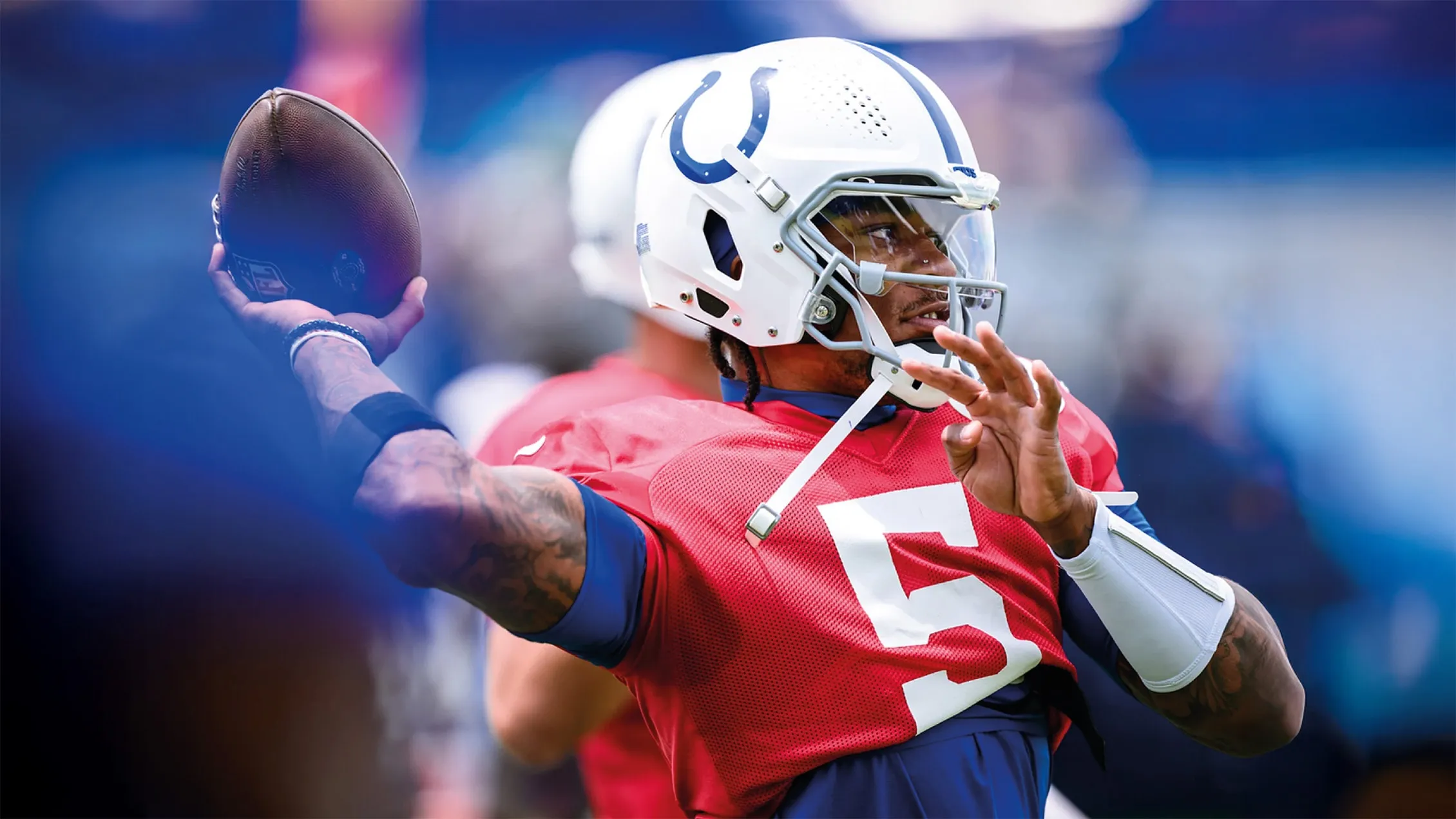 Football player in white helmet with blue horseshoe and red jersey number 5 preparing to throw a football.