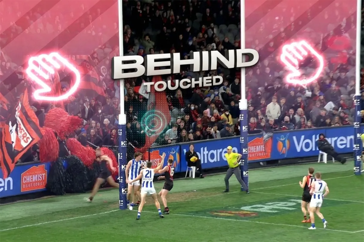 Australian rules football players contest the ball near the goalposts with a crowd in the background and 'BEHIND TOUCHED' text overlay.