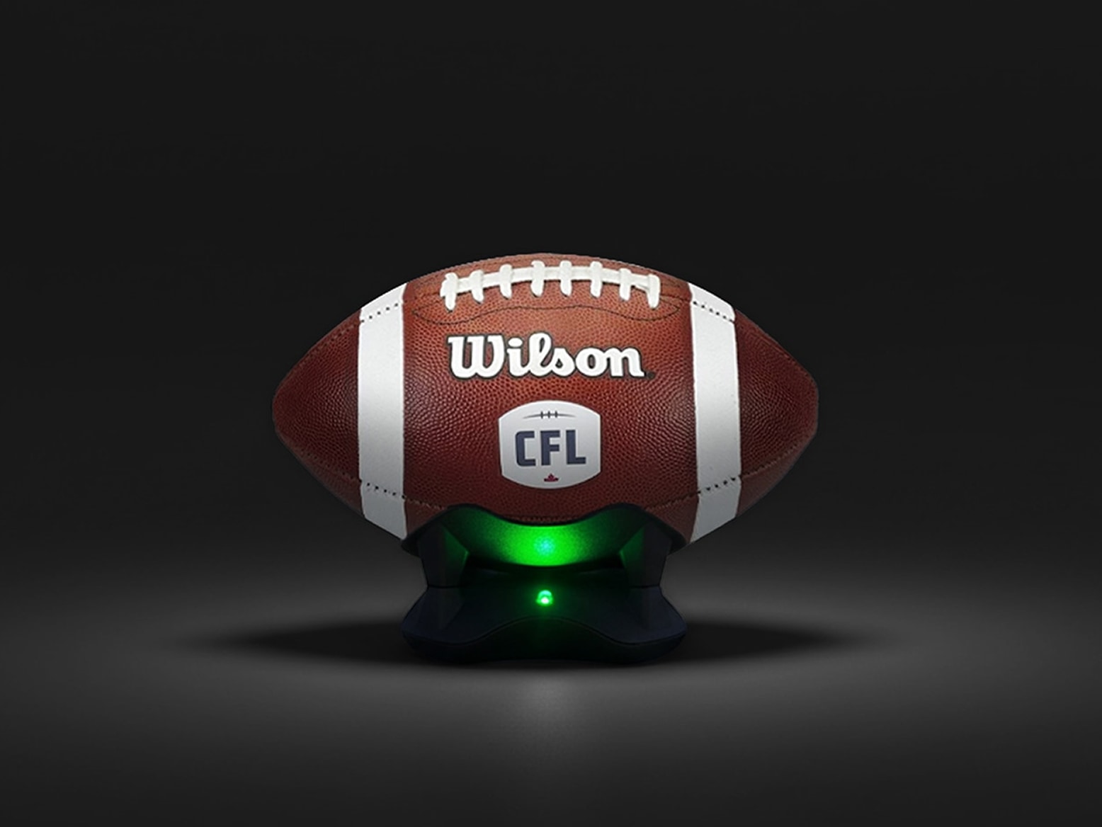 Wilson CFL-branded brown leather football with white stripes placed on a black charging stand emitting green light.