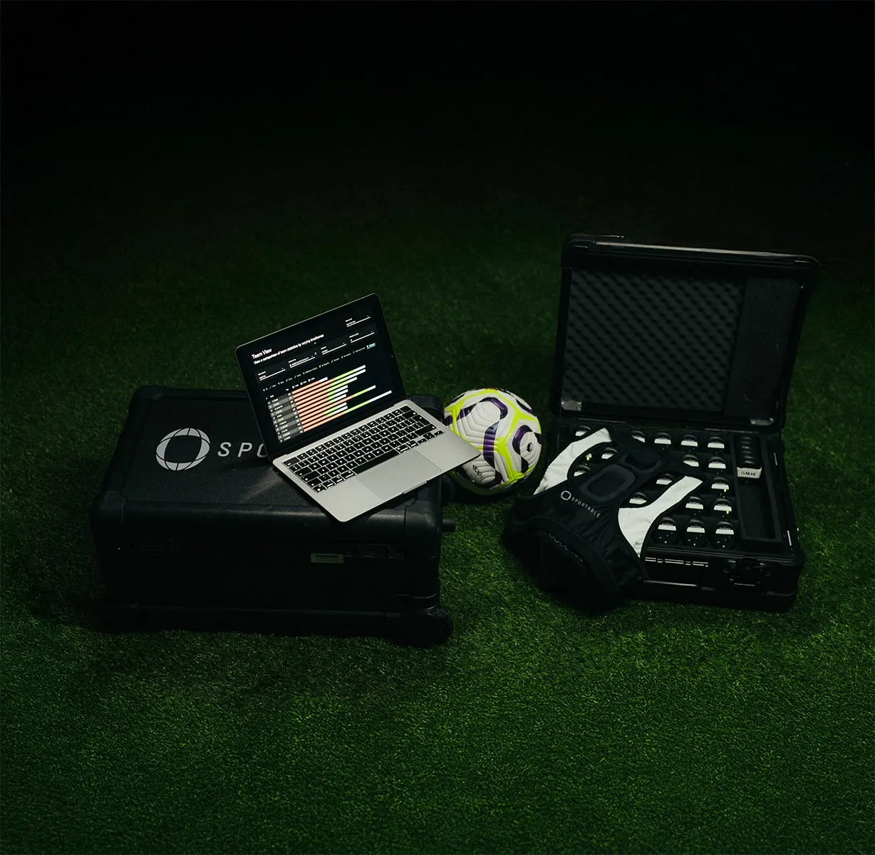 Open laptop displaying a team performance dashboard, a soccer ball, and a black case with wearable sports sensor devices on a grass field.
