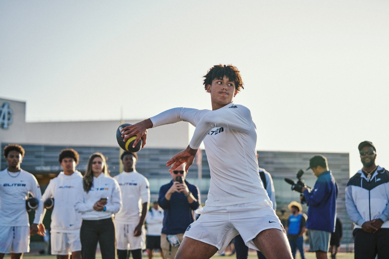 Quarterback winding up to throw at a practice session while staff and players watch