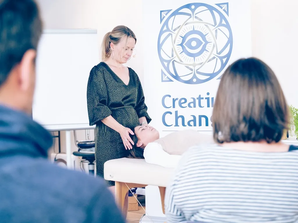 Woman standing and gently holding the head of a person lying on a massage table during a workshop with two attendees watching.