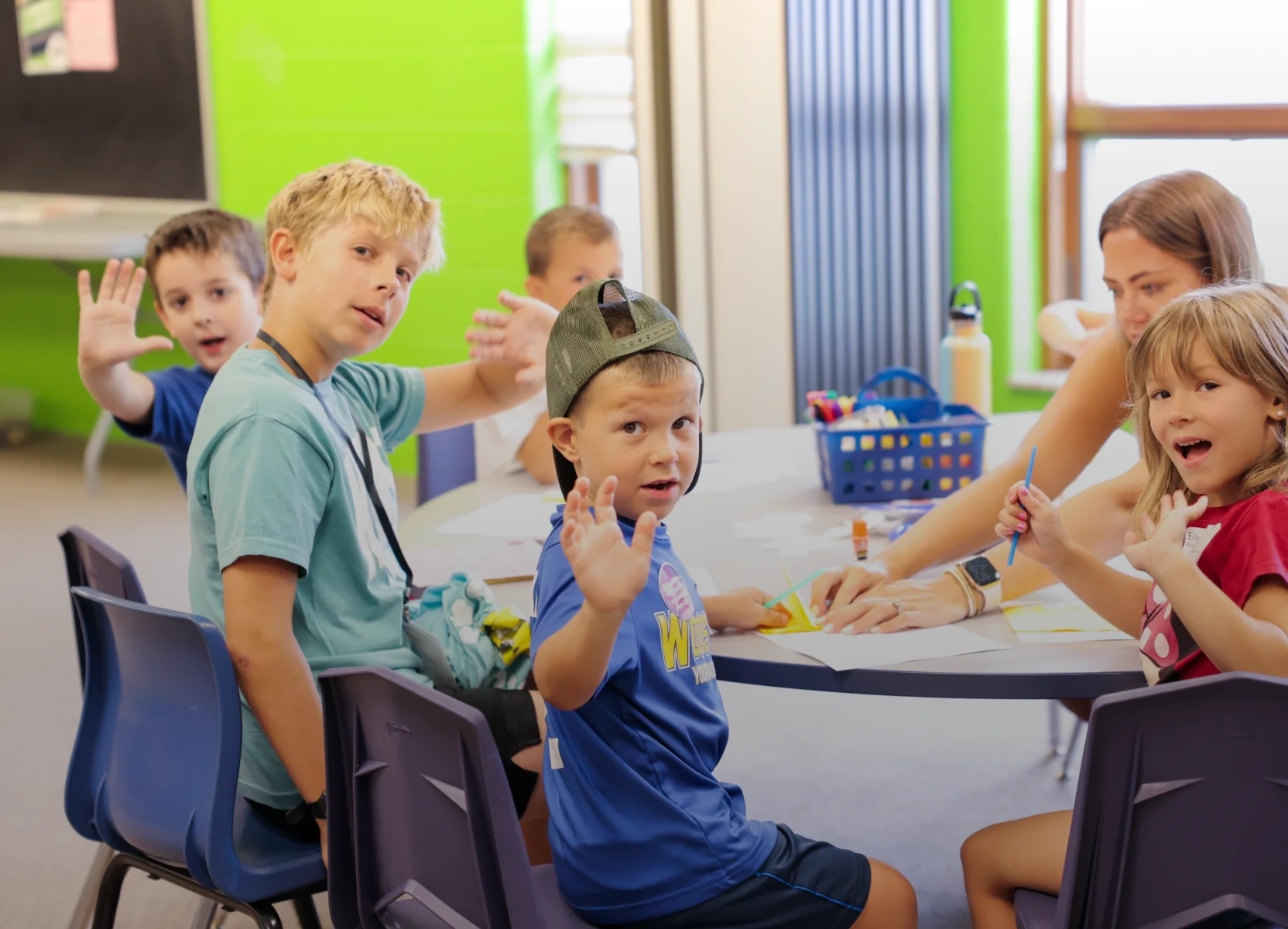 Group of children sitting around a table in a classroom, waving and smiling with a woman helping them with crafts.