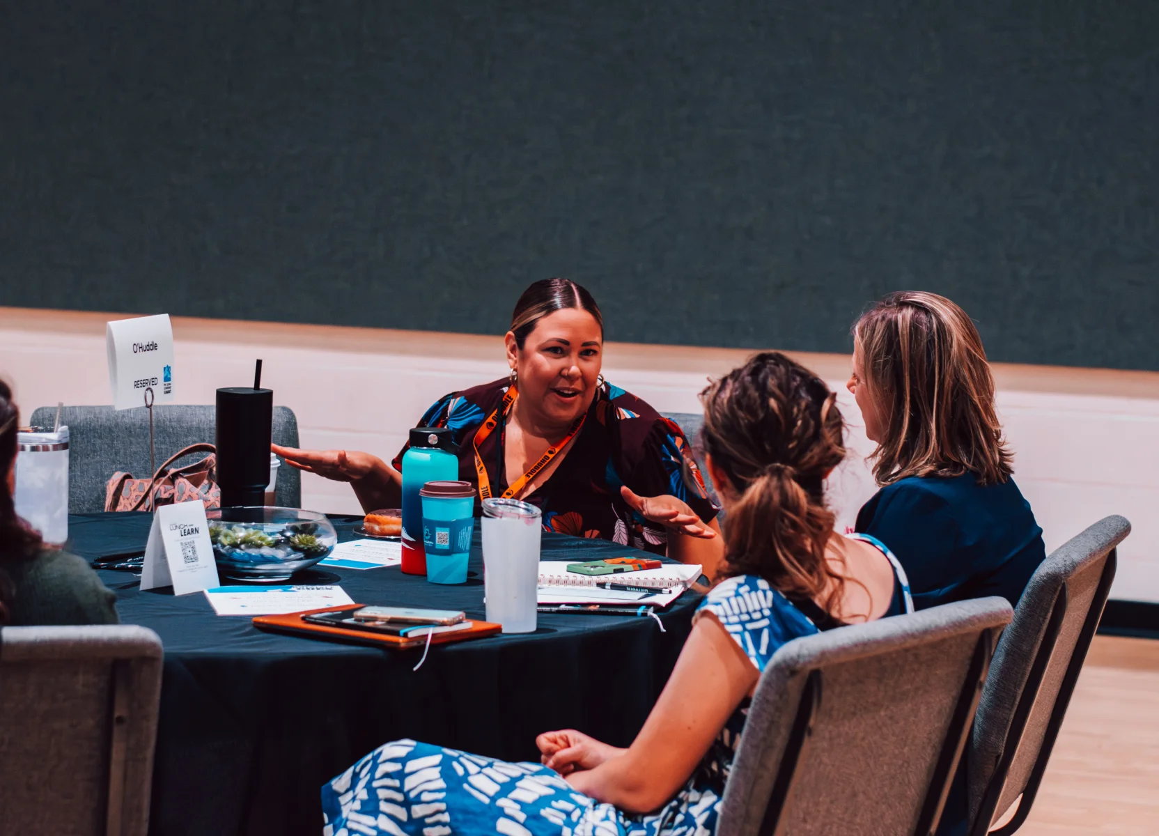 Three women engaged in conversation around a table with drinks, notebooks, and a reserved sign.