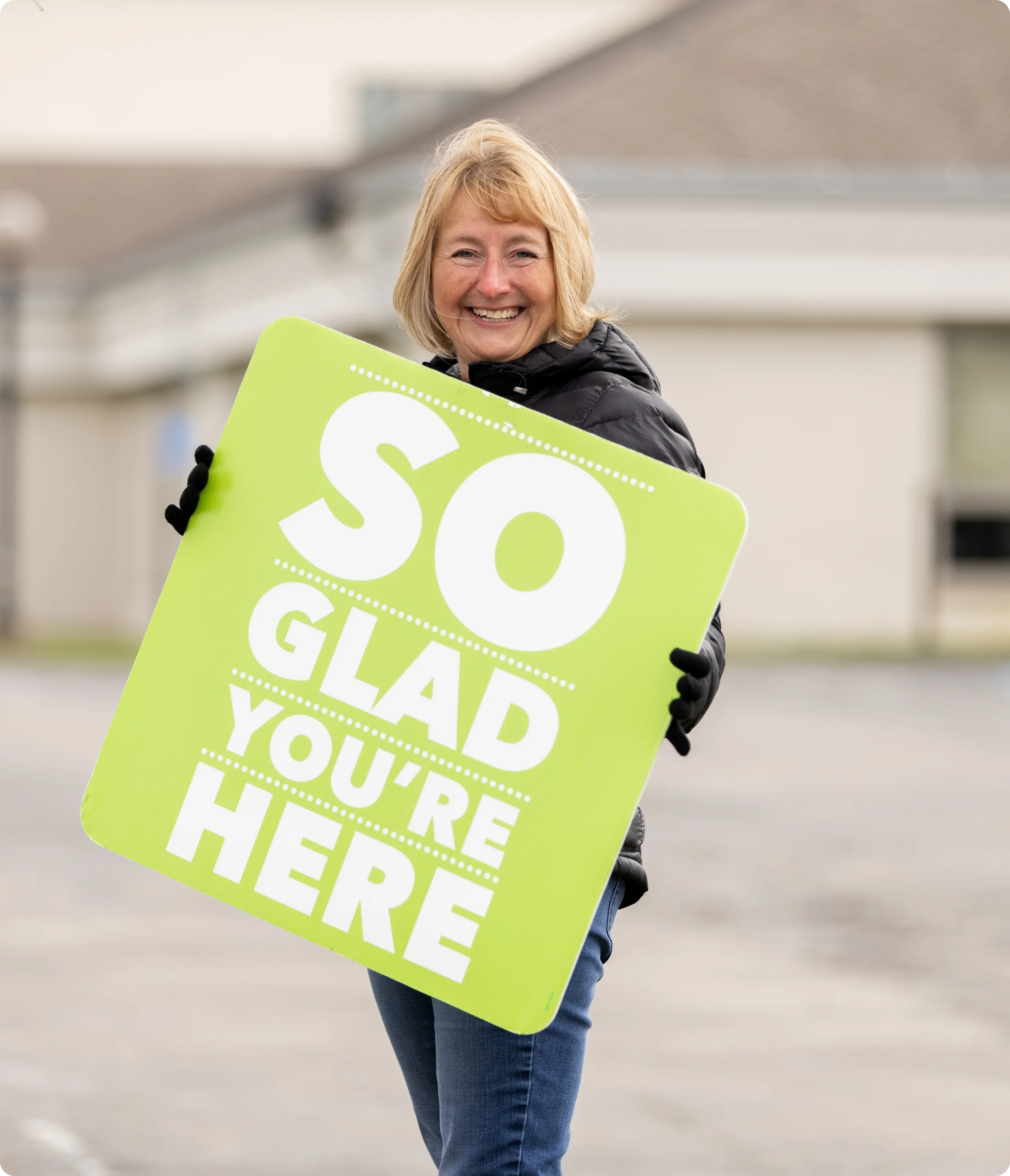 Smiling woman in a black jacket holding a green sign that reads 'So glad you're here'.