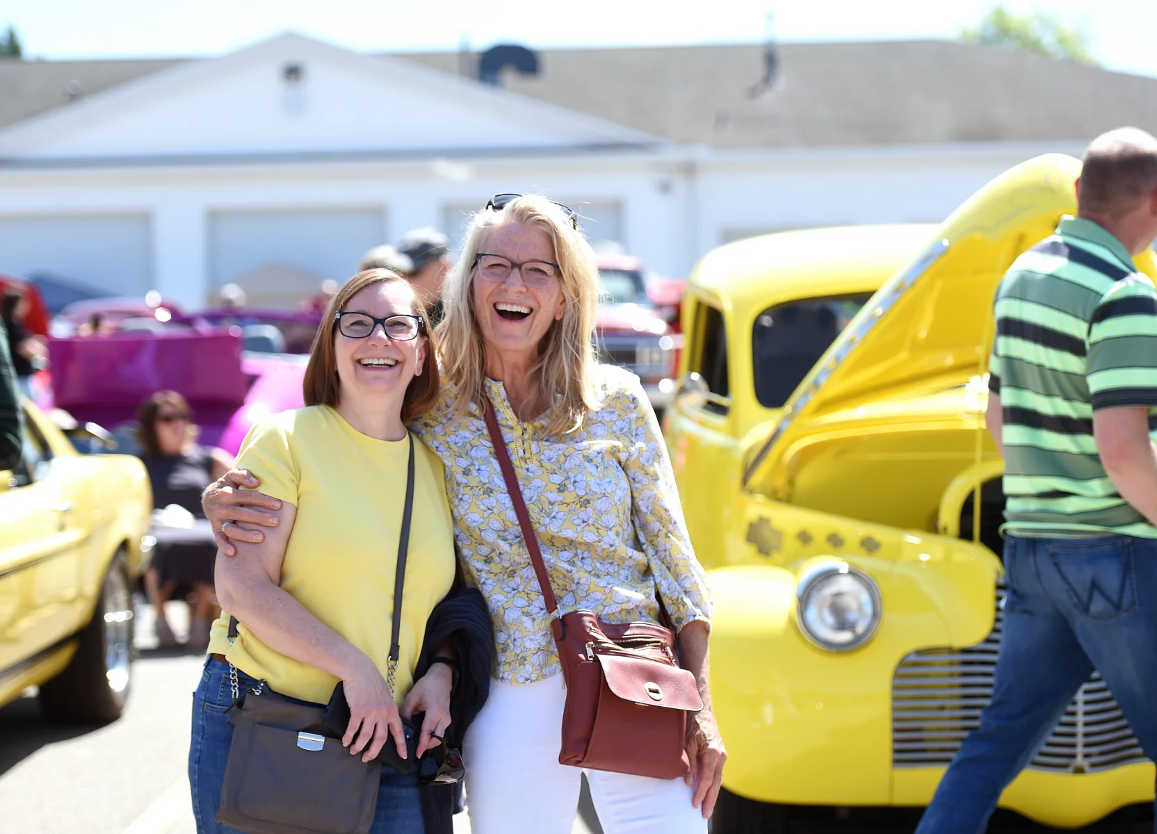 Two smiling women with glasses standing arm in arm in front of a yellow classic car at an outdoor car show.