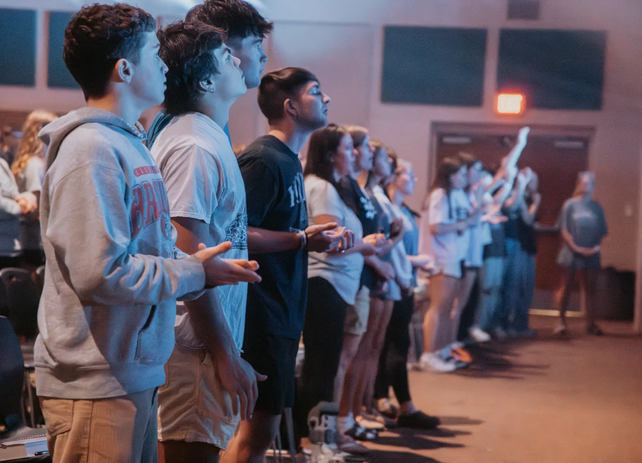 Group of young people standing indoors, focused and engaged, some with hands raised during a gathering.