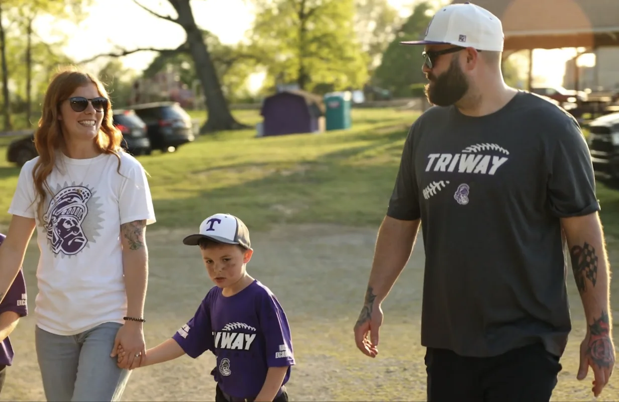 Family of three walking outdoors near a baseball field, woman holding a child's hand, wearing baseball-themed shirts and caps.