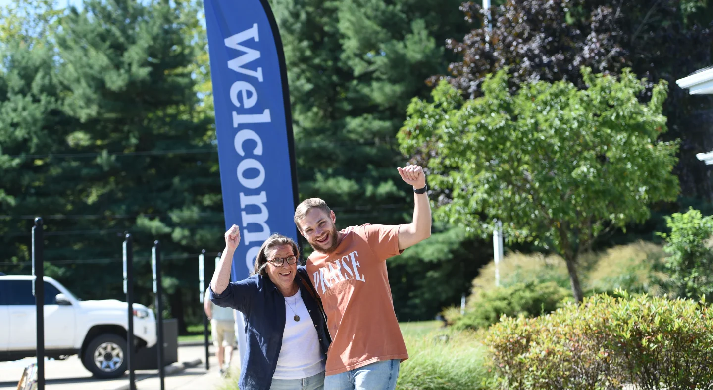 Smiling man and woman waving outdoors beside a blue welcome banner with trees and parked white vehicle in background.