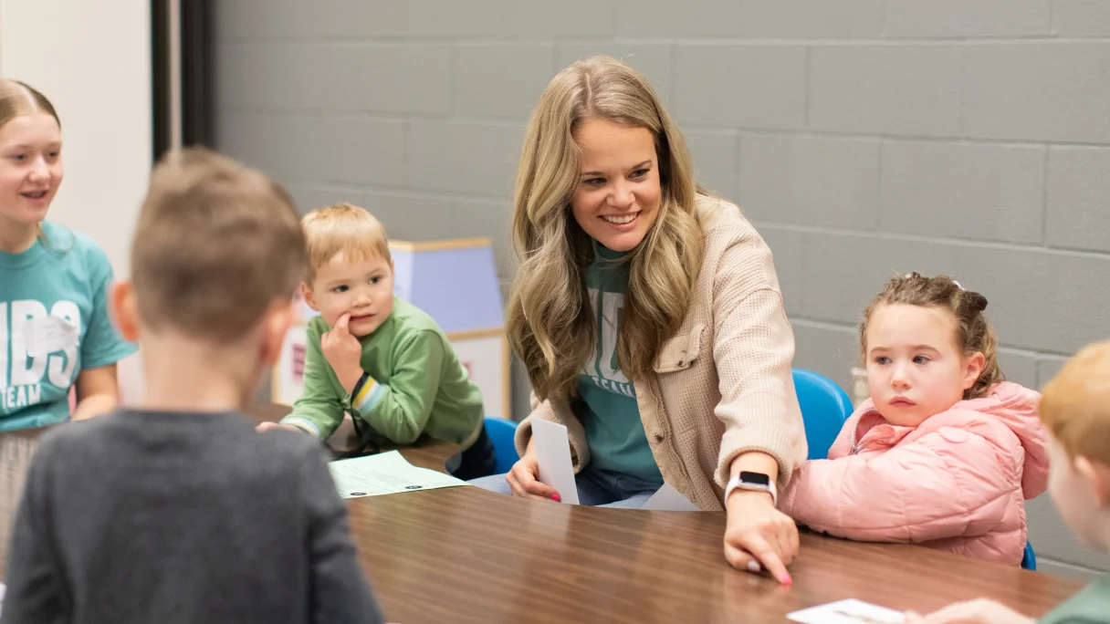 Teacher smiling and engaging with young children seated around a table in a classroom.