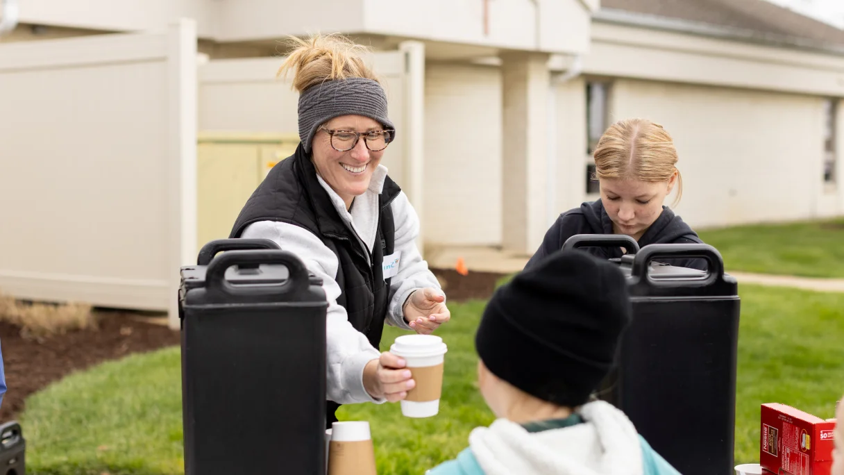 Smiling woman serving a cup of coffee to a child outdoors near a building.