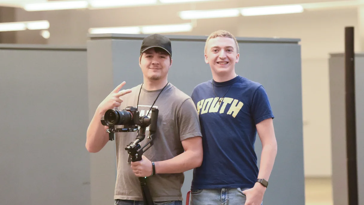 Two young men smiling indoors; one holds a camera stabilizer and makes a peace sign.