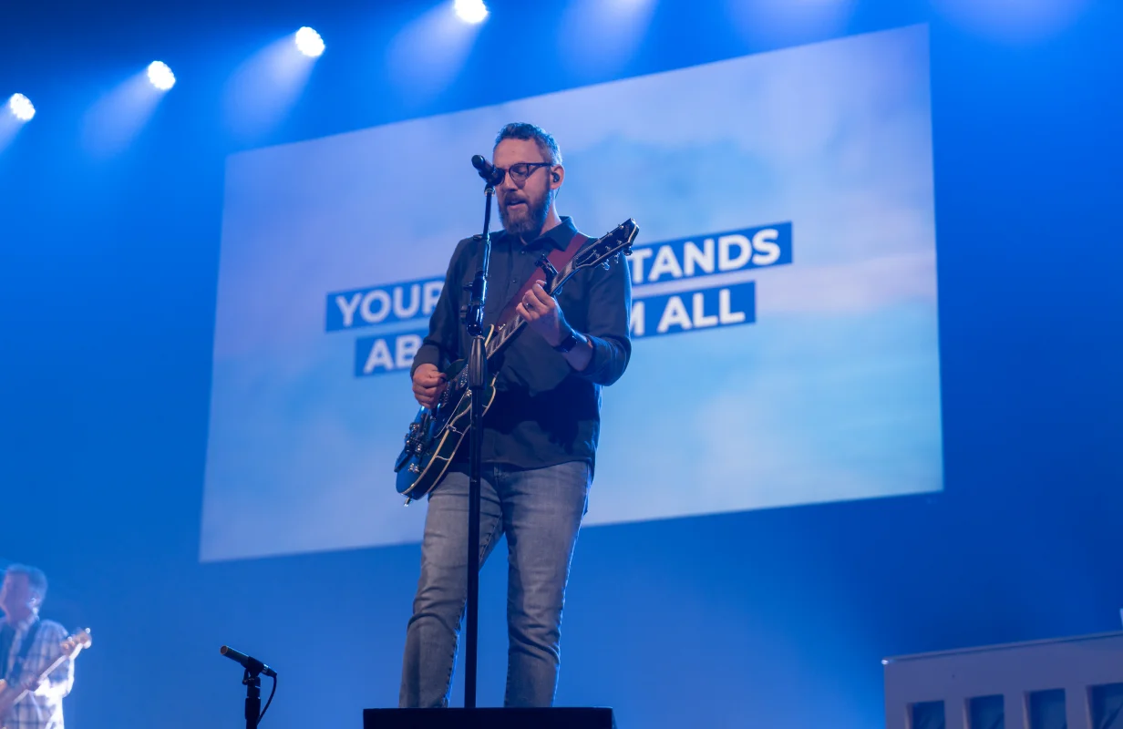 Man singing and playing electric guitar on stage with microphone and blue lighting, a screen behind displays song lyrics.