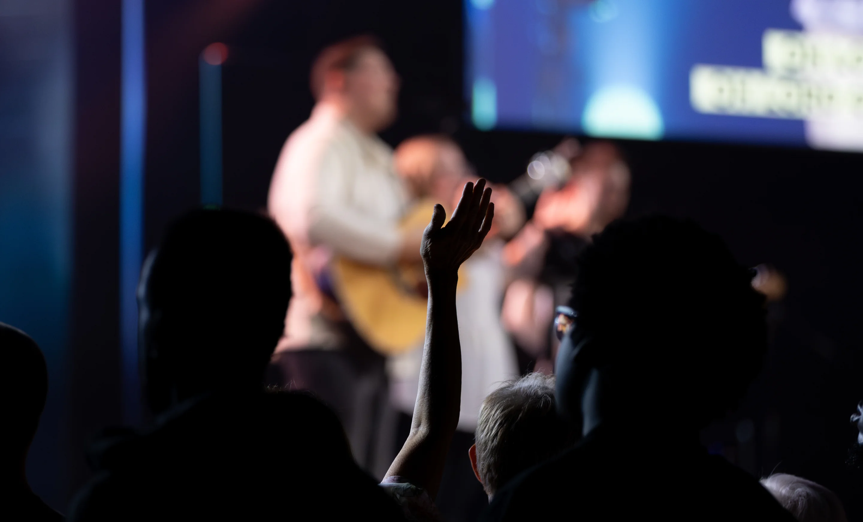 Silhouettes of people with one raised hand during a worship service with a band playing in the background.