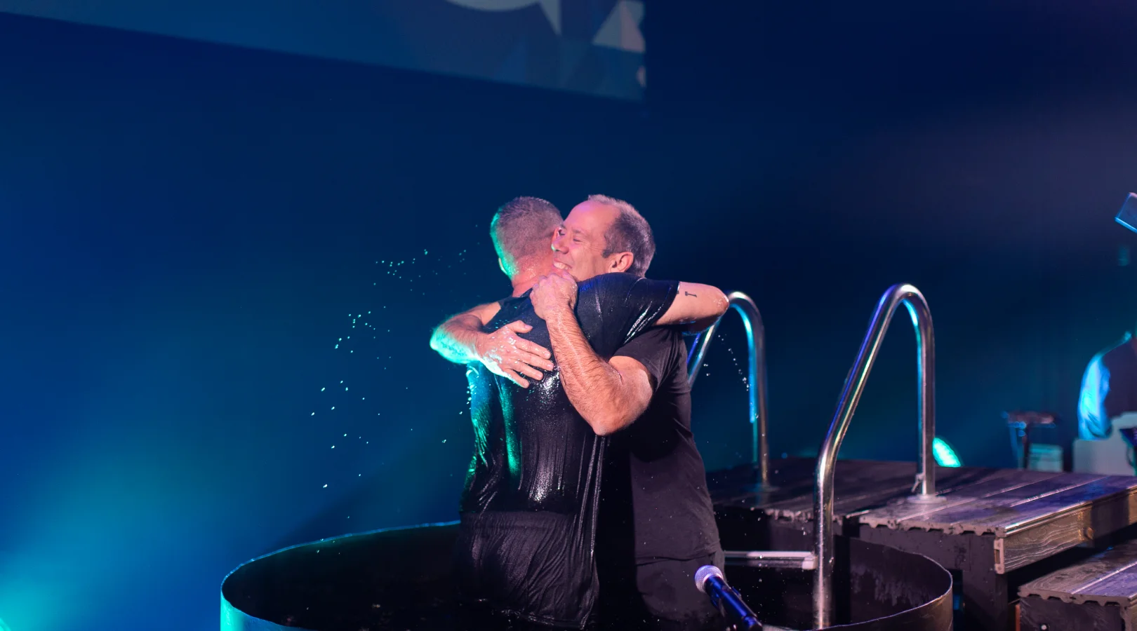 Two men embracing and smiling in a baptismal pool with water droplets around them.