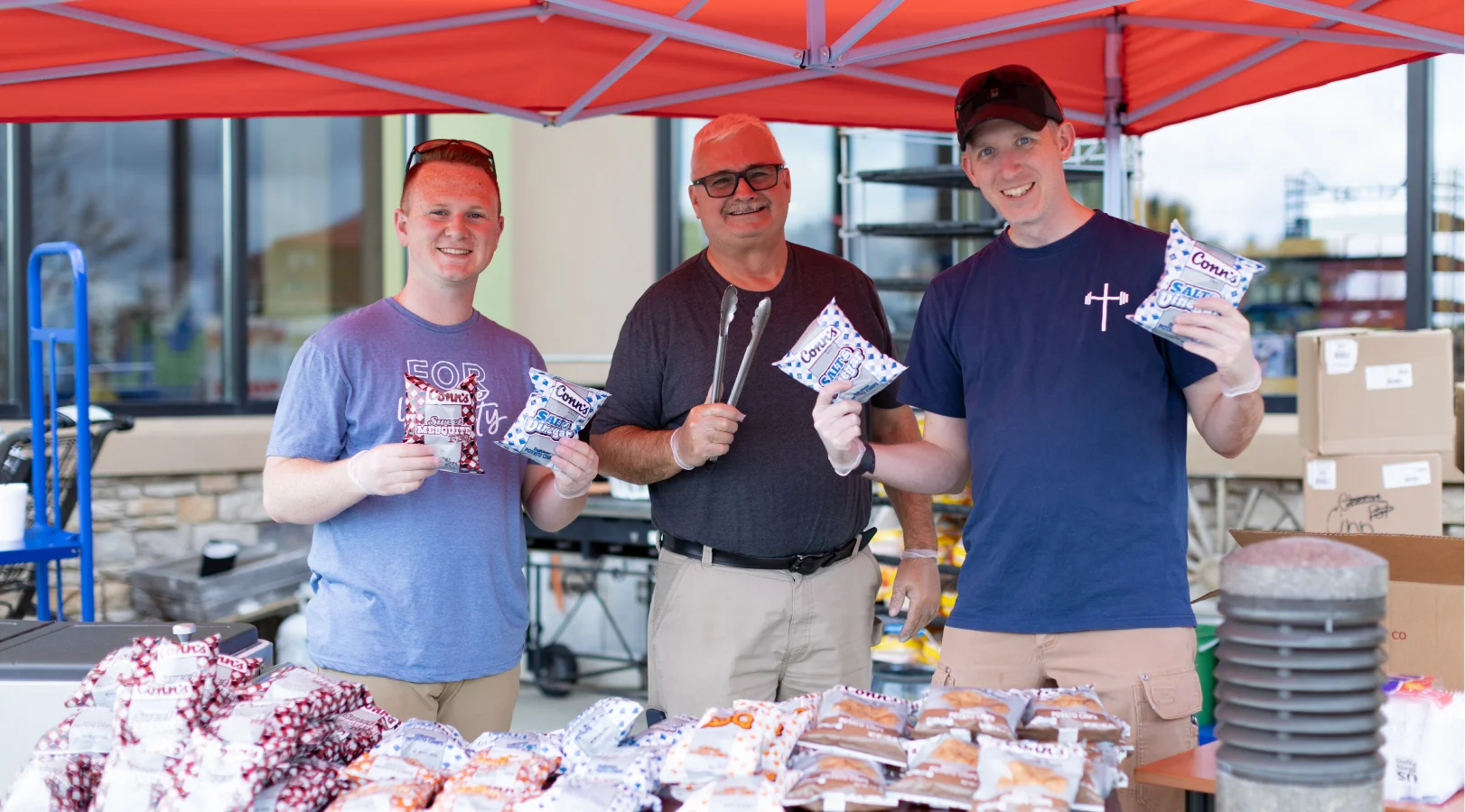 Three volunteers smiling under a red tent holding packets of snacks with more snack bags laid out on a table in front of them.
