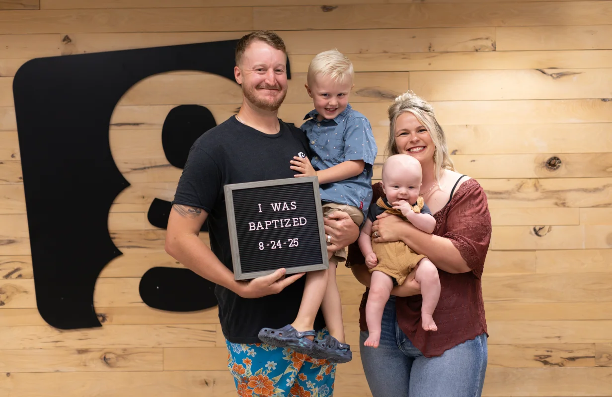 Smiling family of four in front of wood panel wall, father holding a sign reading 'I WAS BAPTIZED 8-24-25'.