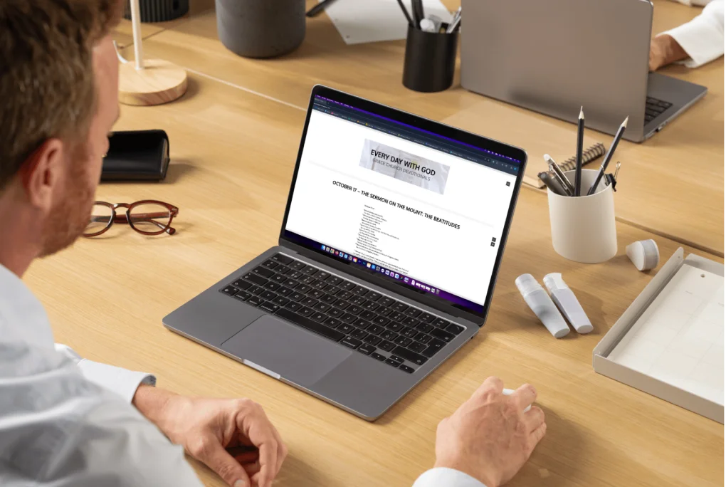 Man seated at a desk, working on a laptop displaying a webpage titled 'Every Day with God' featuring church devotionals.