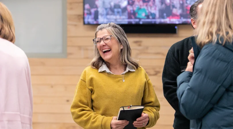 Middle-aged woman in a mustard yellow sweater smiling and holding a book while talking to others in a room with wooden walls.