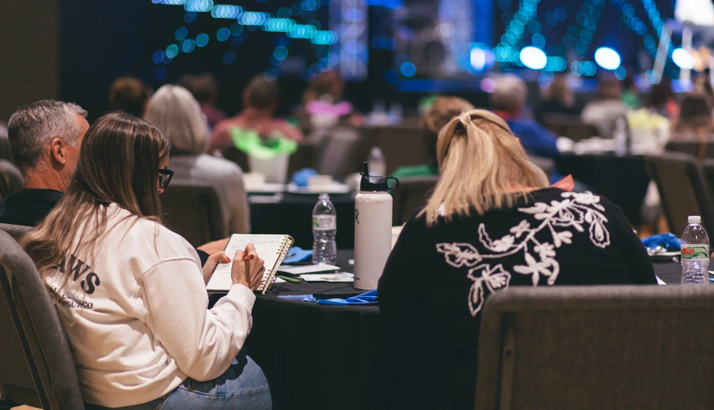 Conference attendees seated at round tables with notebooks and water bottles, taking notes during an event.