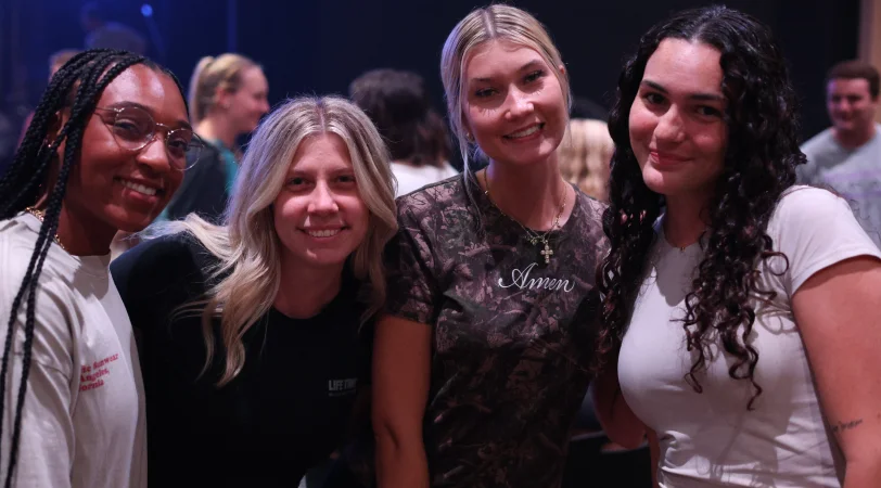 Four smiling young women standing close together indoors, with a blurred crowd in the background.