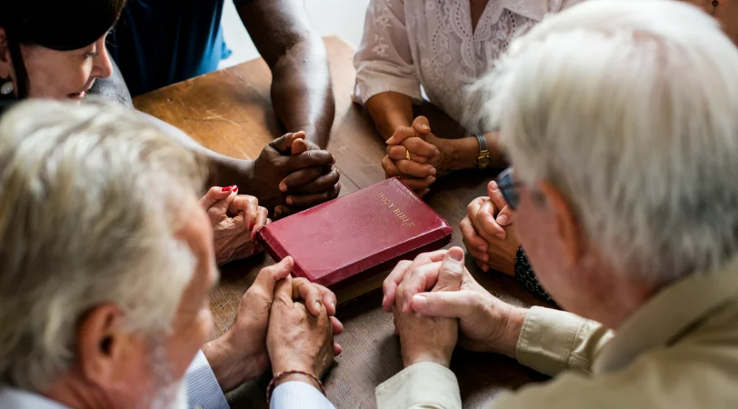 Diverse group of people sitting around a wooden table with a Bible, holding hands in prayer.