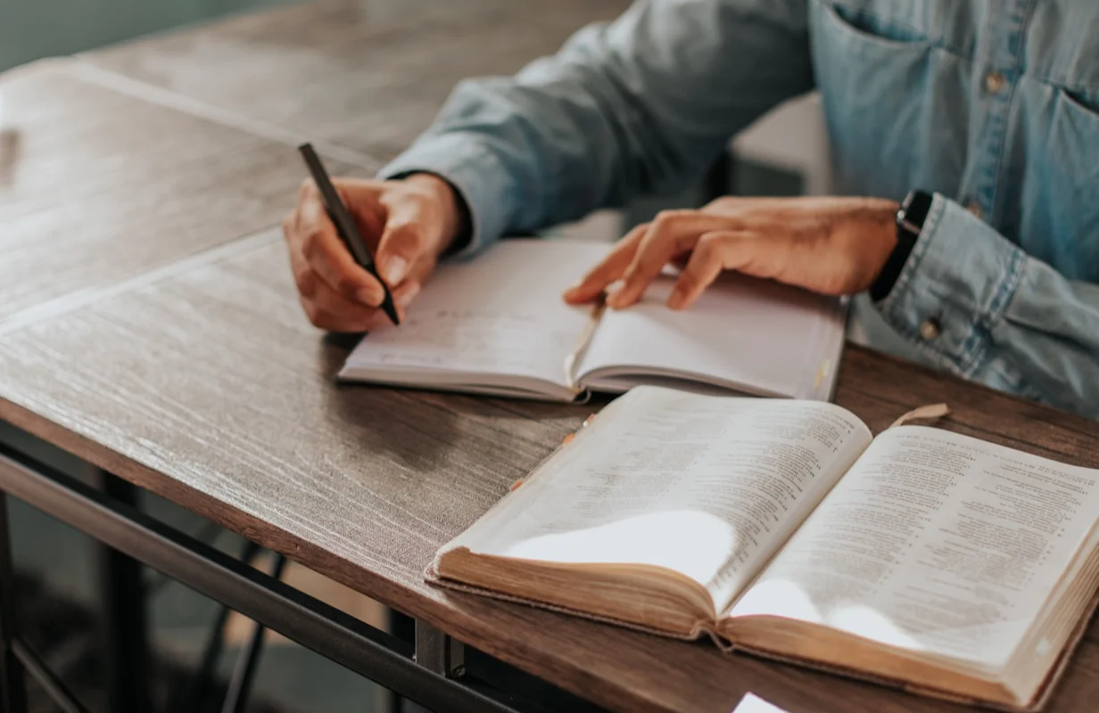 Person writing in a journal with a pen next to an open Bible on a wooden table.