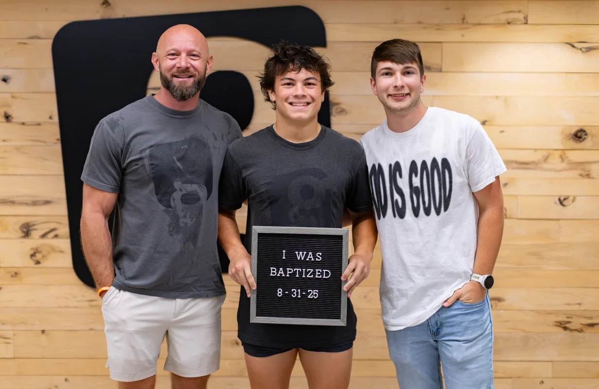 Teen boy holding a sign reading 'I WAS BAPTIZED 8-31-25' standing between two smiling men in a church lobby with a wooden wall background.
