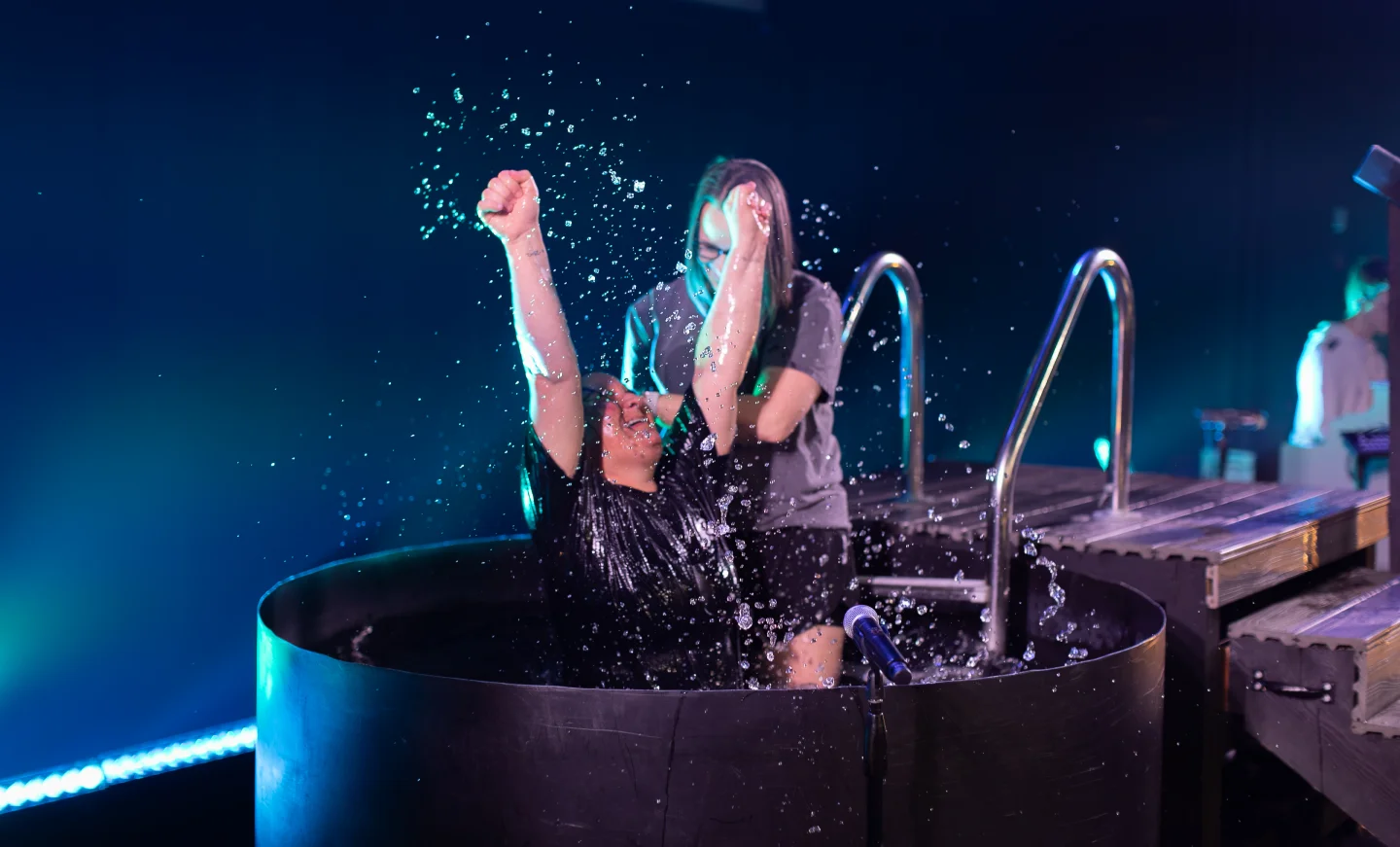 Person raising arms in celebration while emerging from a baptismal pool with another person assisting.