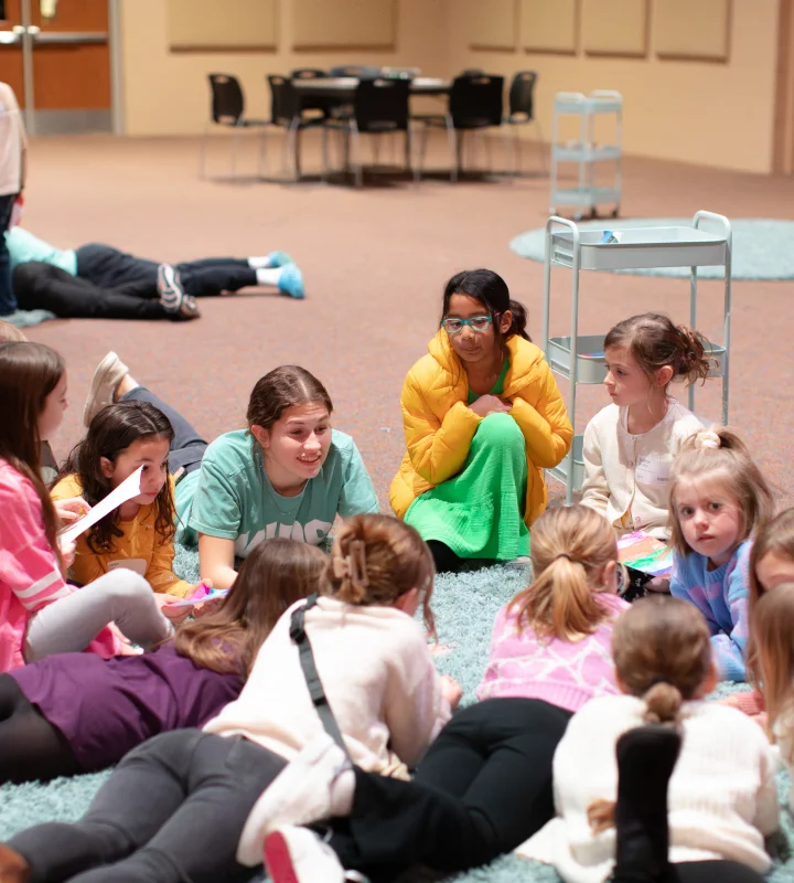 Group of children sitting and lying on a carpet in a circle during a storytime or lesson in a spacious indoor room.