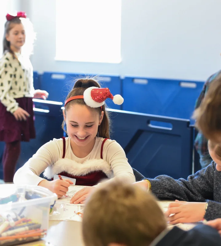 Smiling girl wearing a festive Santa headband coloring at a table with other children in a bright room.