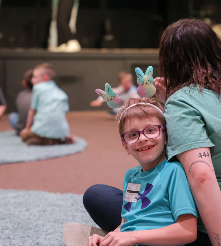 Smiling child wearing glasses and bunny ears headband, sitting next to an adult with a '444' tattoo on their arm, in a group setting with children in the background.