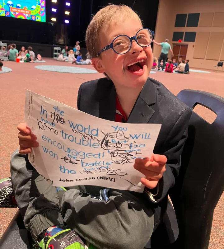 Smiling young boy with glasses, seated and holding a handwritten sign with a Bible verse from John 16:33b (NIRV) at a church event.
