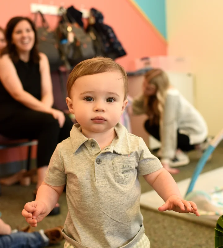 Toddler wearing a grey polo shirt standing indoors with two women sitting and crouching in the background.