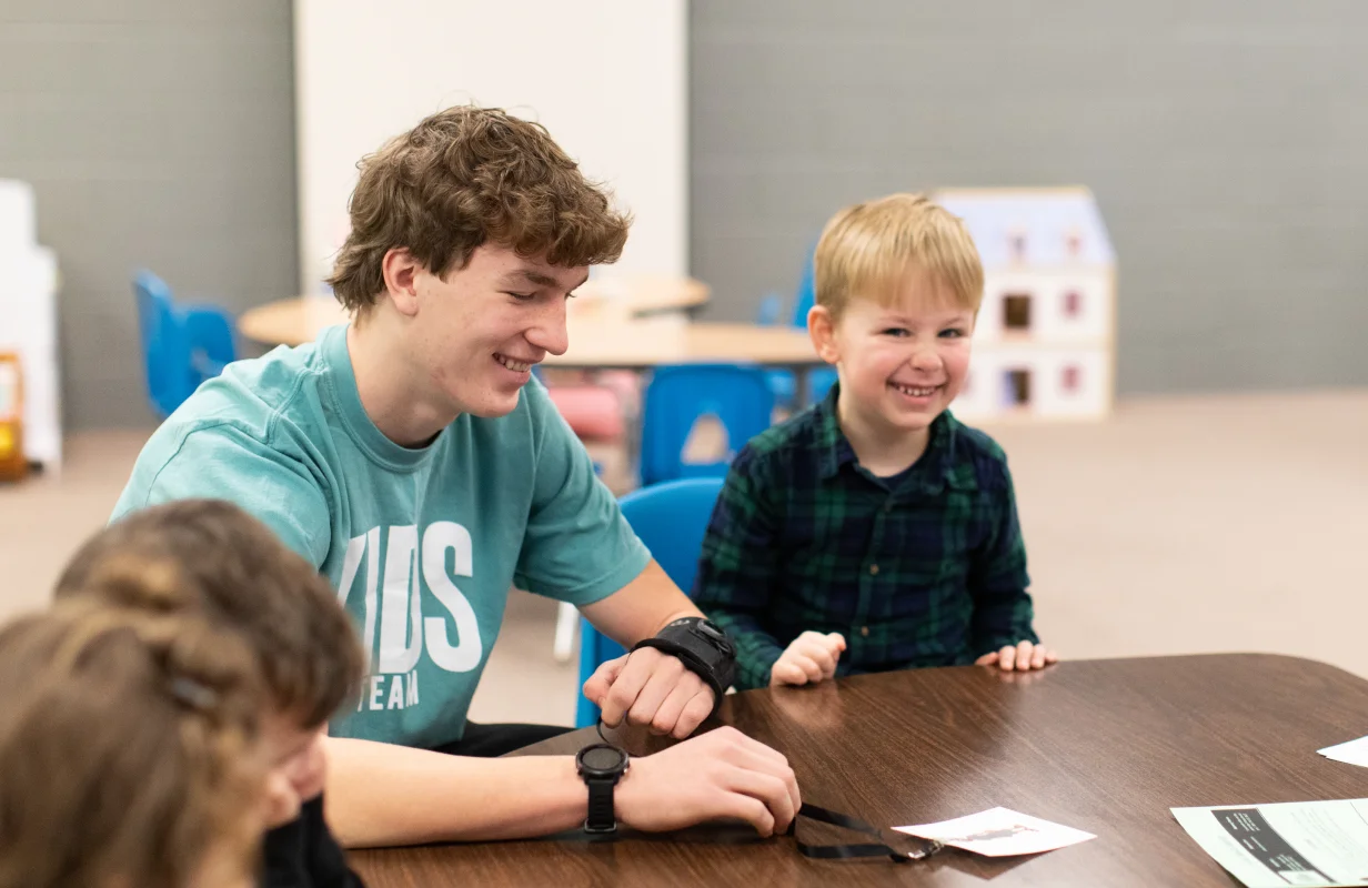 Teen volunteer wearing a teal Kids Team shirt smiling and playing with a young boy at a table.