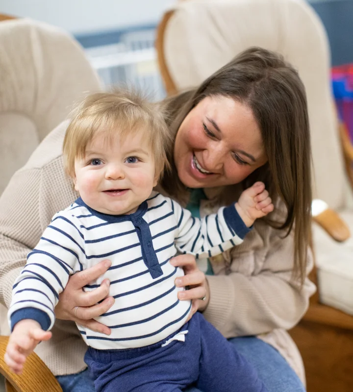 Smiling mother holding a happy toddler dressed in a striped shirt and blue pants indoors.