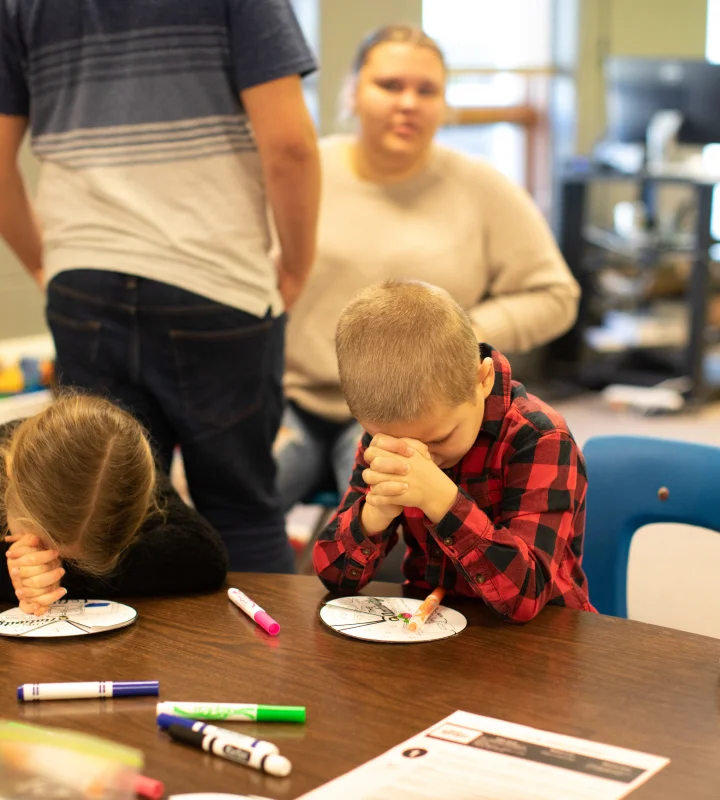 Two children sitting at a table with paper plates and markers, heads bowed in prayer or deep thought.