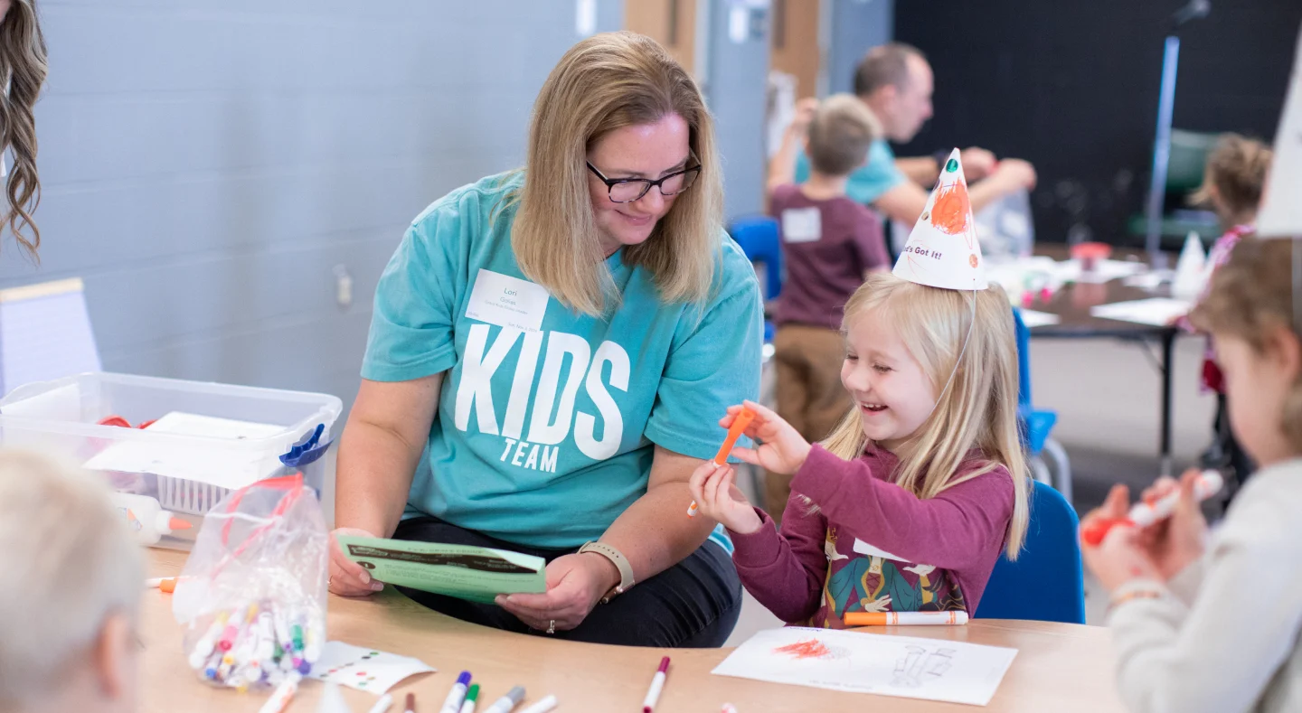 A woman wearing a blue 'Kids Team' shirt helps a young girl with craft activities at a table filled with markers and paper.
