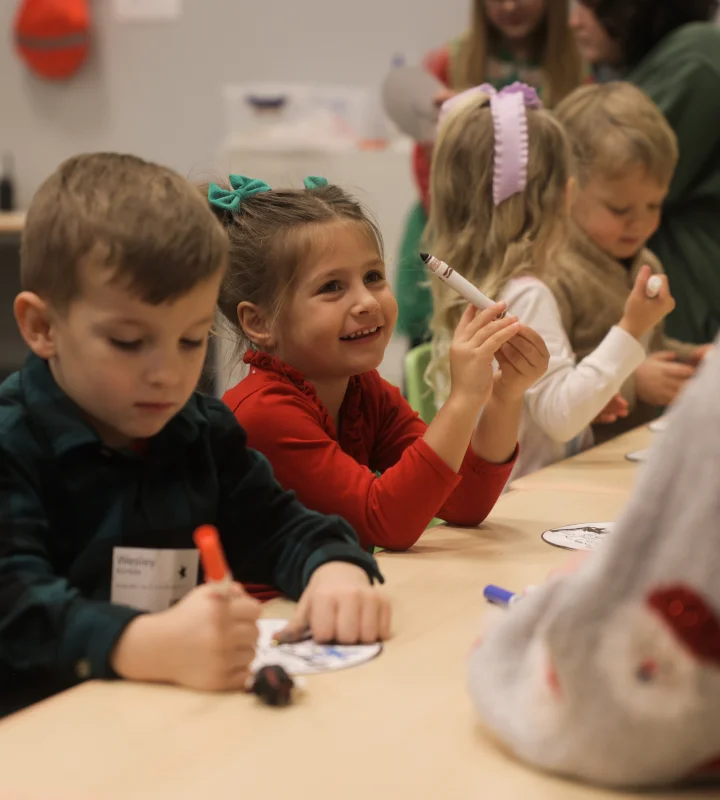 Children sitting at a table in a classroom, coloring and holding markers, with one girl smiling and raising a white marker.