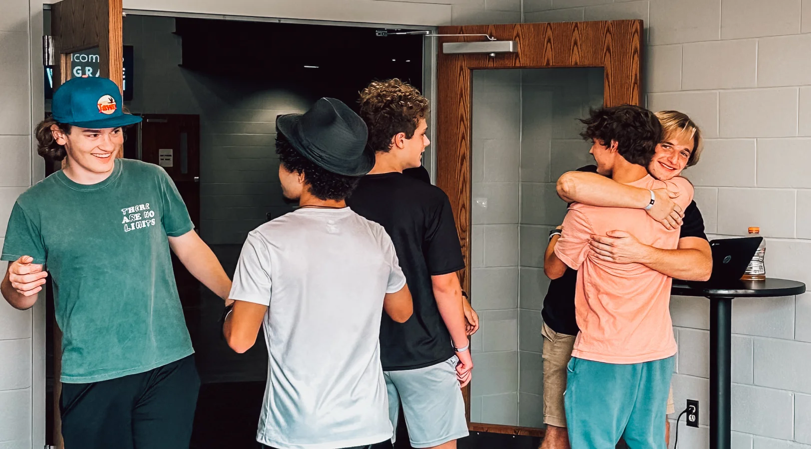 Five teenage boys in a hallway, two hugging and smiling, others standing nearby talking and smiling.