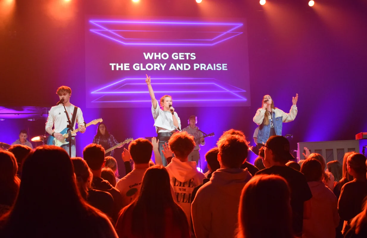 Youth band performing on stage with purple lighting and a screen behind displaying the text 'Who gets the glory and praise' while audience members raise their hands.