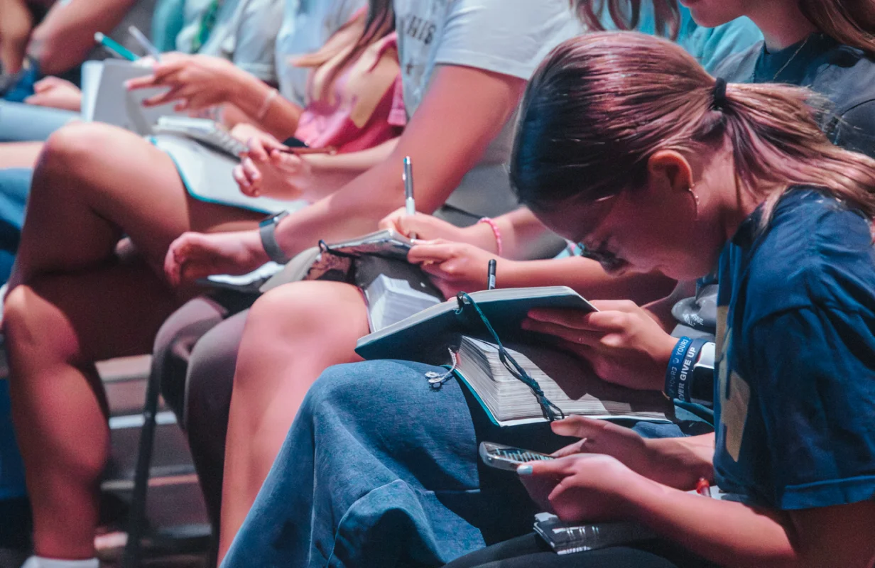 Group of young people sitting closely, writing notes on notebooks and holding pens during a youth service.