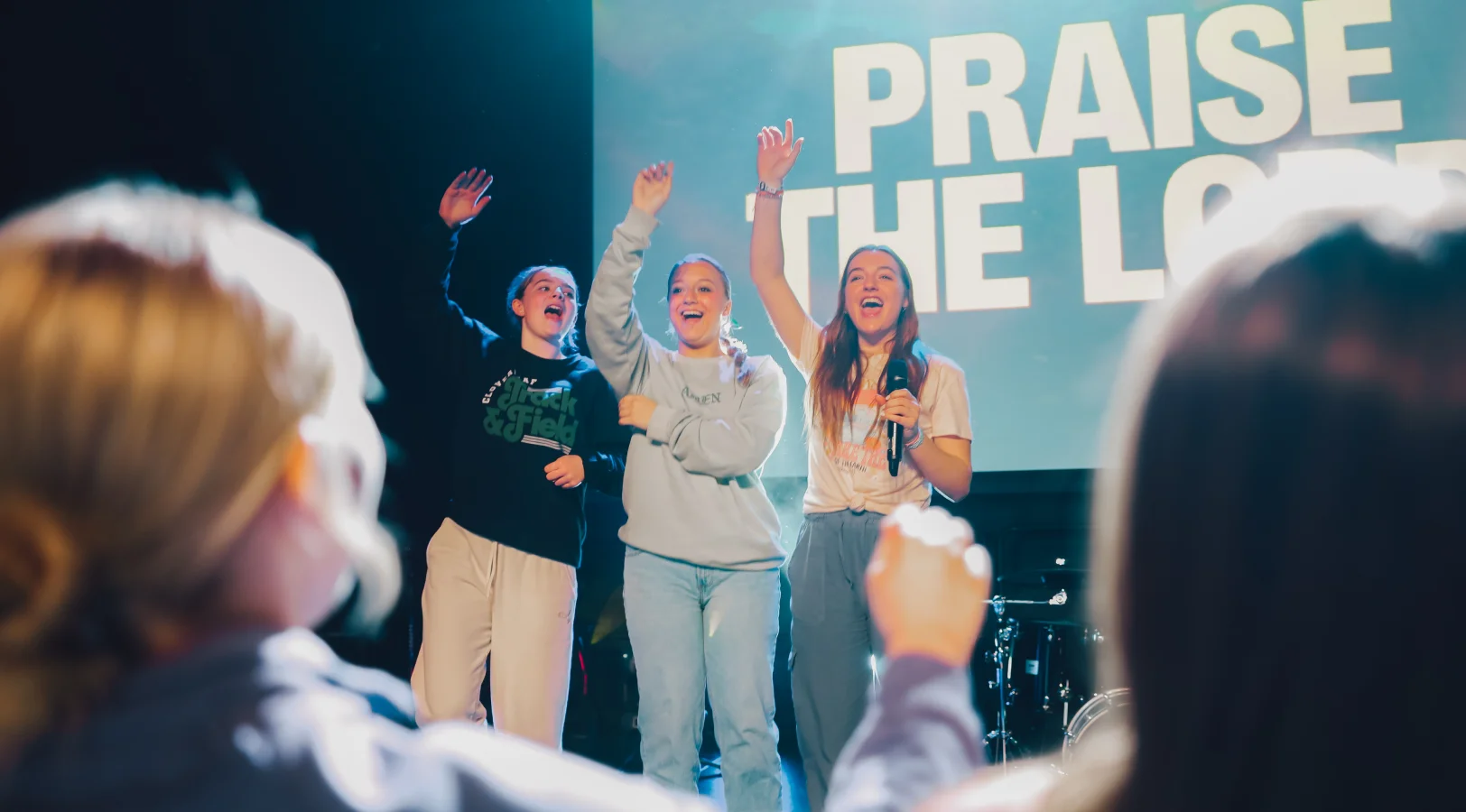 Three young women on stage raising their hands and singing during a praise worship service, with an audience in the foreground and large text that reads 'PRAISE THE LORD' in the background.