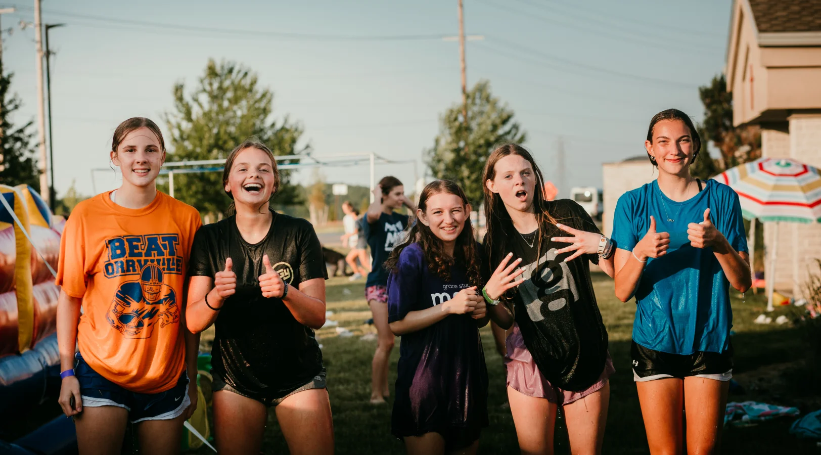 Five smiling girls outdoors in casual athletic clothing during a sunny event, two giving thumbs up.