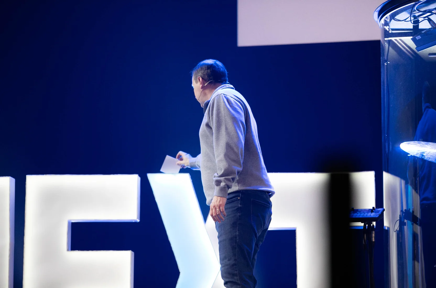 Man in a gray sweater and jeans holding a note card, standing on stage with large illuminated letters in the background.